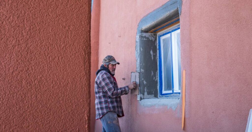 A worker wearing a flannel shirt uses a tool to scrape away paint from a window frame on an adobe building.