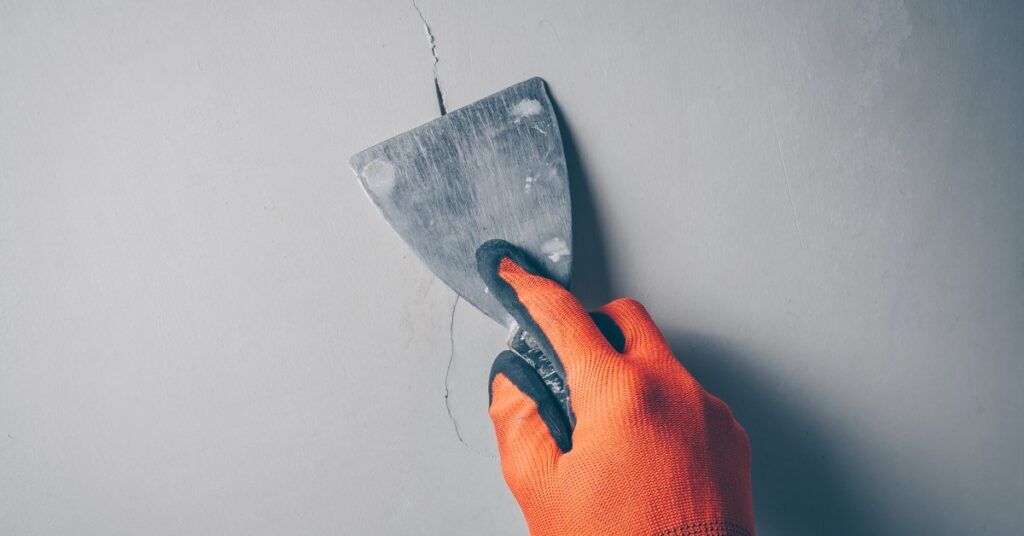 A worker wears a glove and uses a trowel to repair the crack in a wall. The wall is white and incredibly bare.