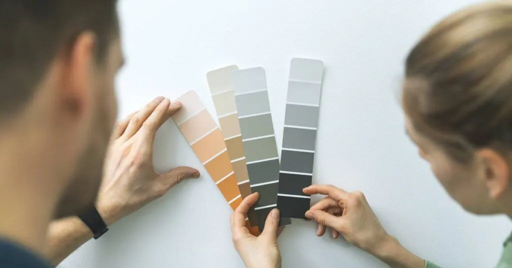 A man and a woman hold up paint samples against a wall. The shades include orange, brown, and gray.