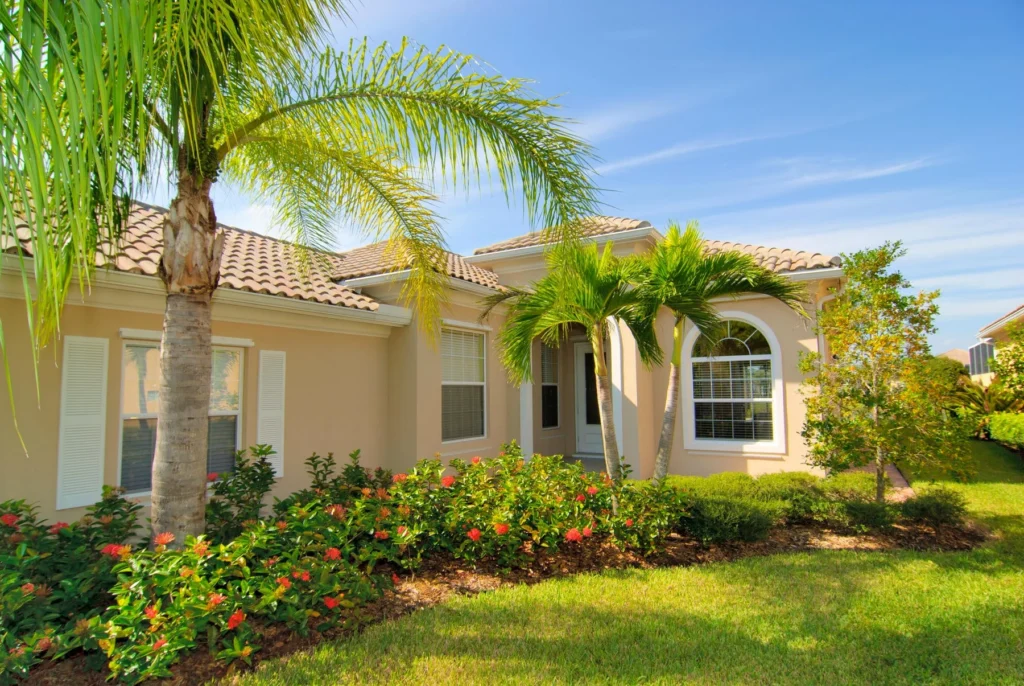 A beige single-story house with white shutters is surrounded by lush landscaping, including palm trees, green bushes, and flowering plants, under a clear blue sky.