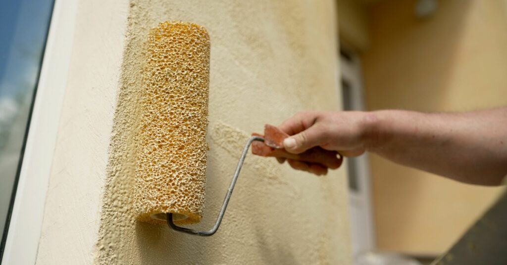 A worker uses a roller brush to put yellow paint on the stucco exterior wall of a home next to a window.