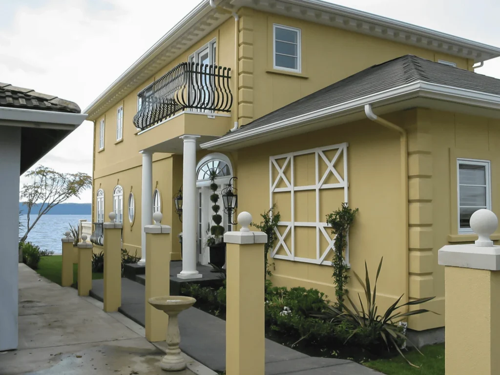 A yellow two-story house with white trim, decorative window frames, a balcony with black railings, white columns at the entrance, and a view of water and sky in the background.