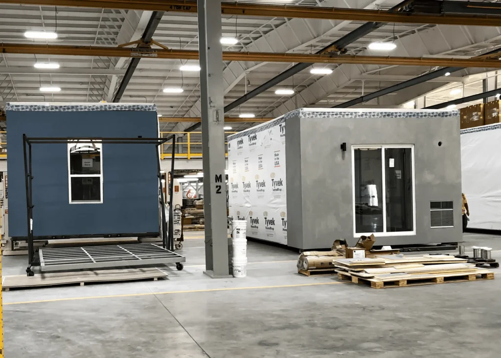 Two prefabricated modular home sections under construction in a large indoor factory; one section is blue with a window, the other is gray with sliding glass doors and construction materials nearby.