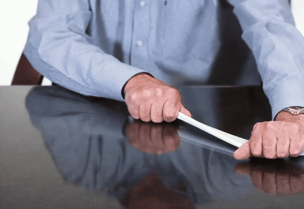 A person in a blue shirt sits at a table, holding a white object with both hands, appearing to twist or bend it. Only the person’s torso, arms, and hands are visible.