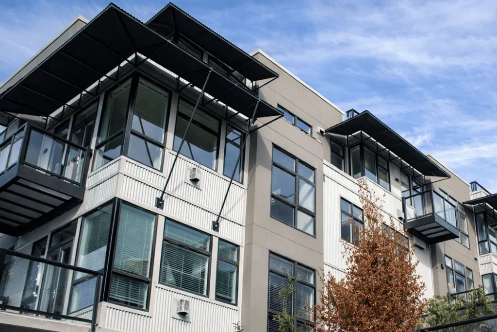 A modern apartment building with large glass windows and balconies, featuring metal and concrete elements, set against a blue sky with some clouds. A small tree with brown leaves is visible in the foreground.