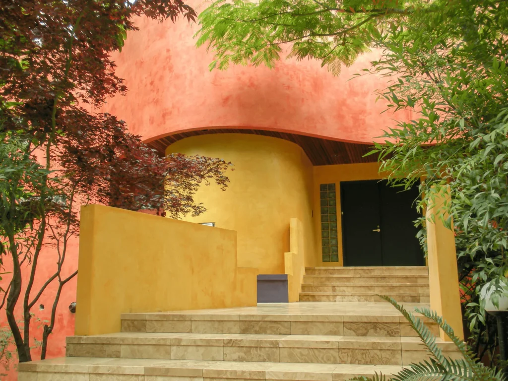 Staircase leading to a modern house with curved yellow and orange stucco walls, surrounded by lush green plants and trees.