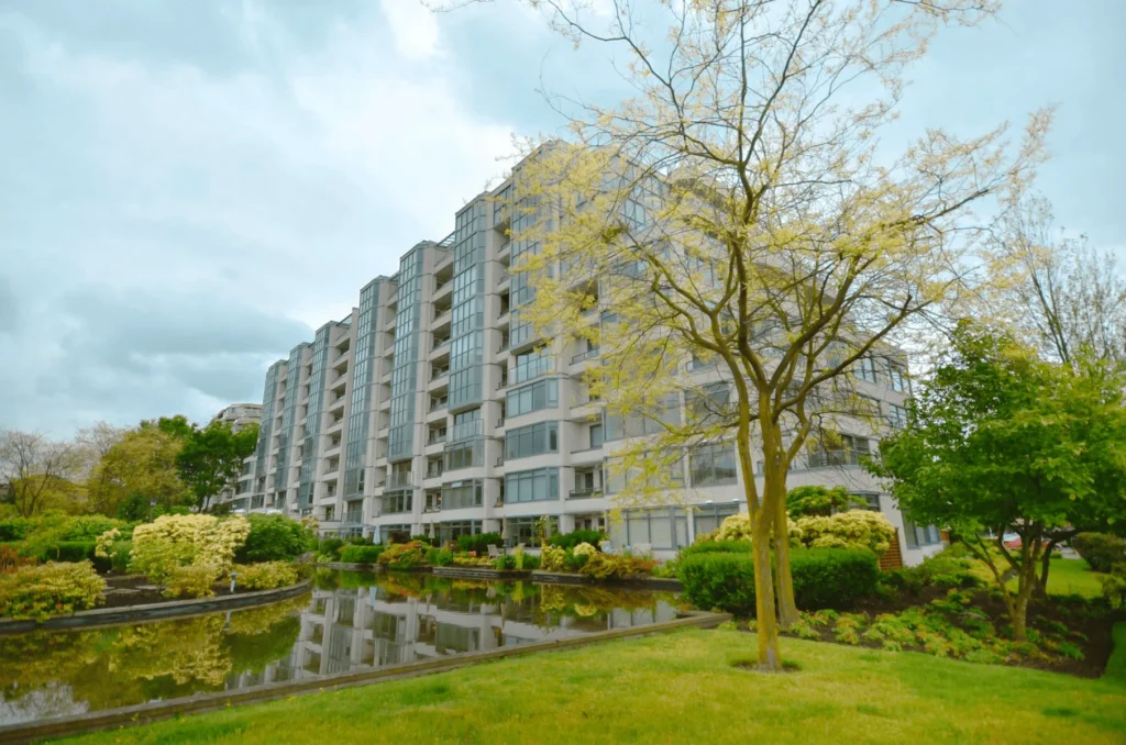 A modern multi-story apartment building with large windows is reflected in a pond, surrounded by green lawns, shrubs, and trees under a cloudy sky.