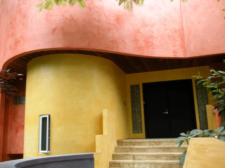 A modern house entrance with curved walls painted in yellow and orange tones, tan steps leading to a black double door, and green plants in the foreground.