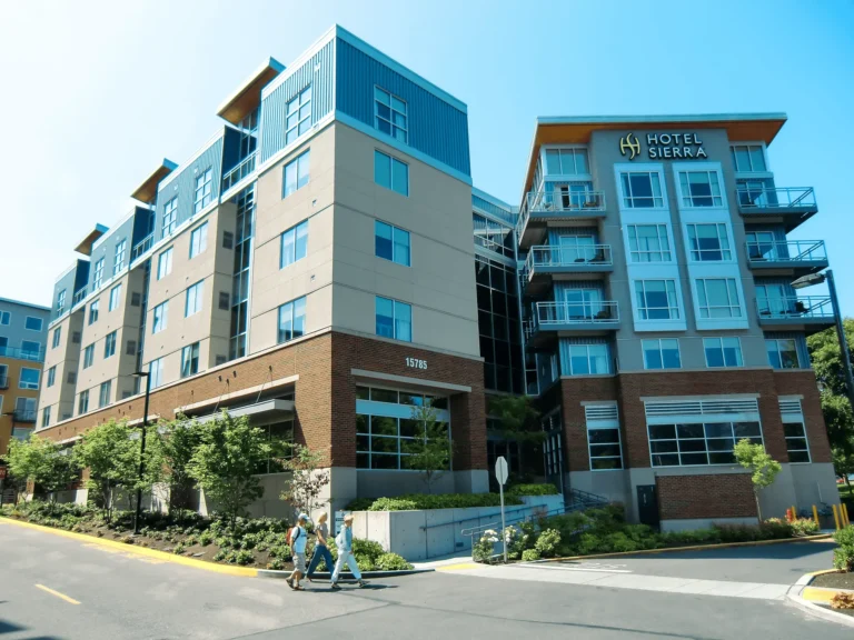Two people walk past a modern multi-story building complex with large windows and balconies; one building has a “Hotel Sierra” sign on its exterior. Trees and landscaping line the street in front.