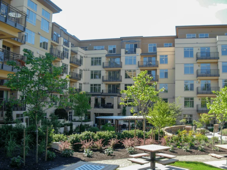 A modern multi-story apartment building with balconies overlooks a landscaped courtyard featuring green trees, shrubs, walking paths, and outdoor seating areas, including a table with a chessboard.