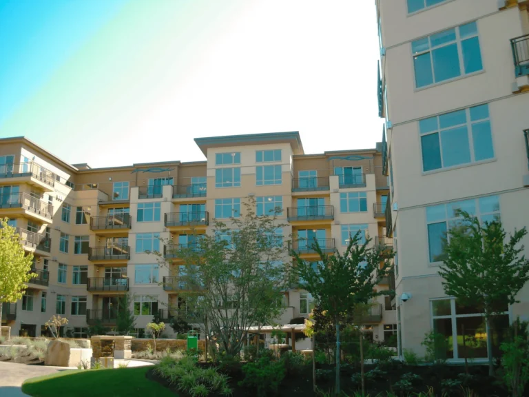 A modern apartment complex with multiple stories, balconies, and large windows, surrounded by landscaped greenery and trees under a bright sky.