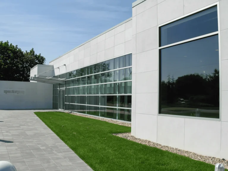 Modern white building with large glass windows, a paved walkway, and a neatly trimmed green lawn on a sunny day. Trees and a blue sky are visible in the background.