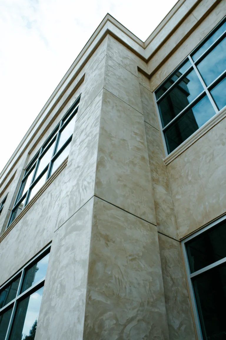 Upward view of a modern building corner with beige stucco walls and large, rectangular windows reflecting the sky. The architecture features clean lines and a minimalist design.