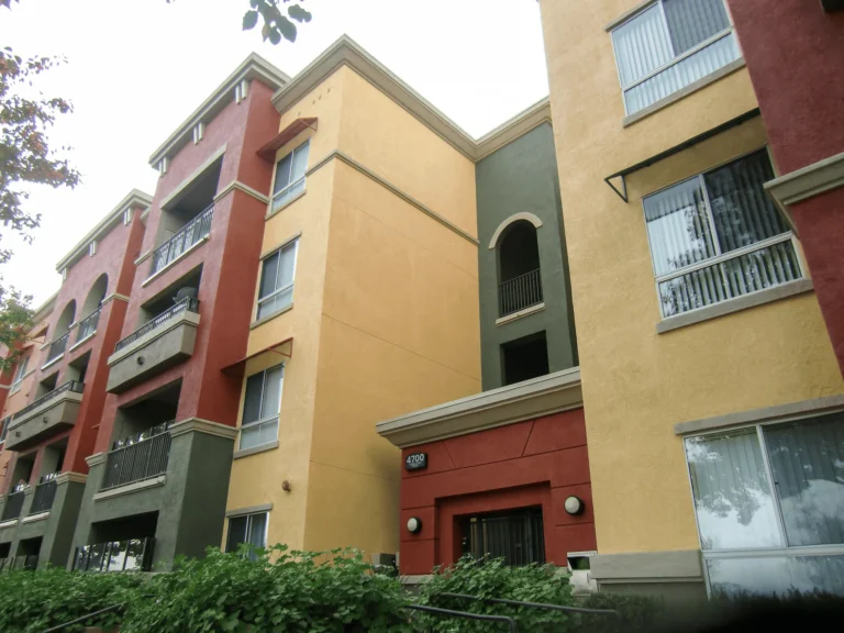 A multi-story apartment building with yellow, red, and green exterior walls, large windows, and balconies, surrounded by greenery and trees.
