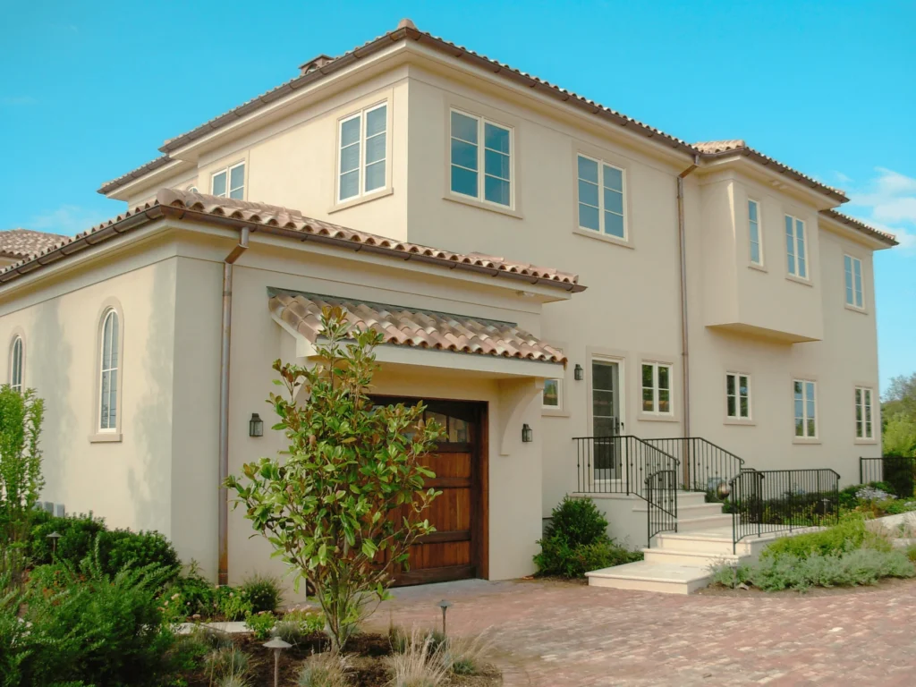 A two-story beige house with a tiled roof, wooden garage door, and multiple windows. There are stairs leading to the front door, surrounded by landscaped shrubs and brick driveway under a clear blue sky.