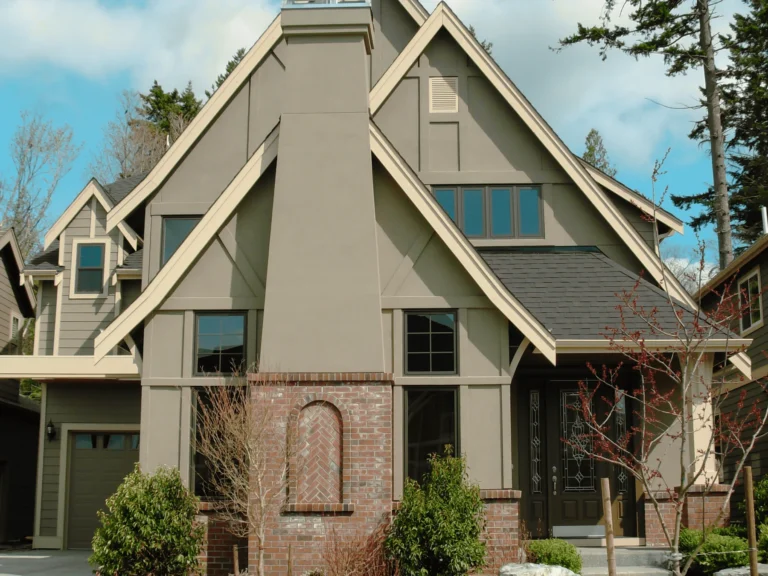 A modern two-story house with taupe siding, black trim, multiple steep gables, large windows, and a brick accent on the front facade, surrounded by small trees and shrubs.