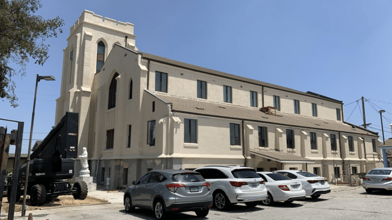A large, light-colored building with tall windows and a tower stands in bright sunlight. Several cars are parked in front, and construction equipment is nearby, indicating renovation work.