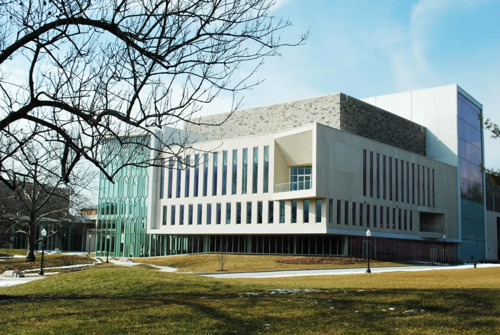 Modern academic building with large windows, stone accents, and a mix of glass and white facade, set on a grassy campus with bare trees and pathways under a partly cloudy sky.