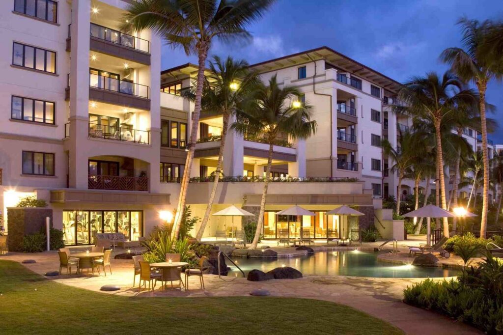 Luxury resort at dusk with palm trees, a lit swimming pool, poolside tables and chairs, and multi-story stucco buildings with balconies glowing with warm lights. The cloudy blue sky enhances the relaxing atmosphere.