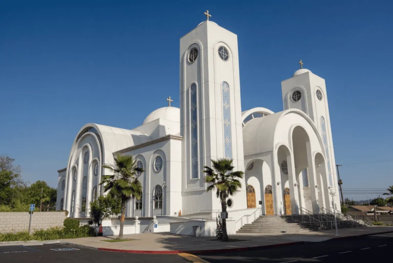 A large white church with domes, arched windows, and tall towers topped with crosses, featuring prominent columns and wooden doors, with palm trees and a clear blue sky in the background.
