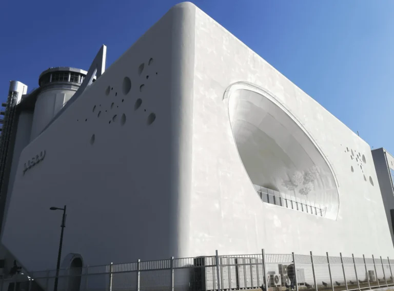 A modern white building with a smooth, curved facade, featuring circular cutouts and a large oval window. The structure is surrounded by a metal fence, and the sky above is clear and blue.