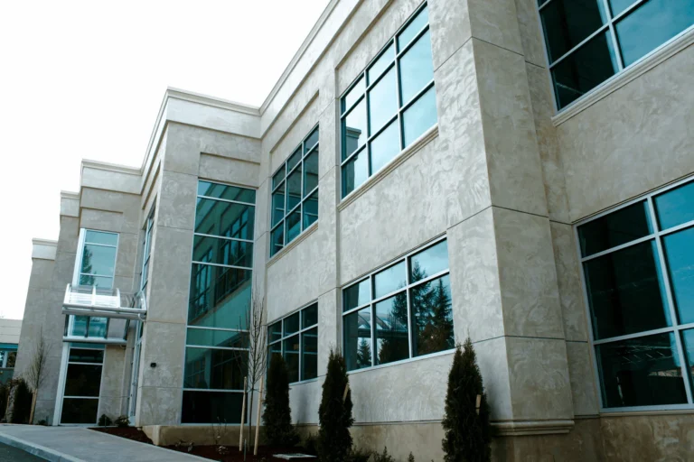 Modern office building with large blue-tinted windows, beige stone exterior, and minimal landscaping including small trees and shrubs near the entrance.