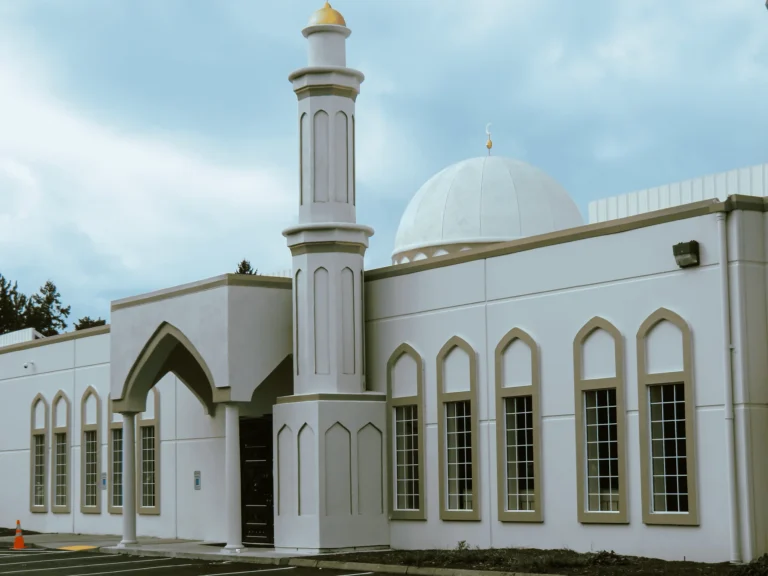 A white mosque exterior with tall arched windows, a prominent minaret, and a large dome, set against a cloudy sky.