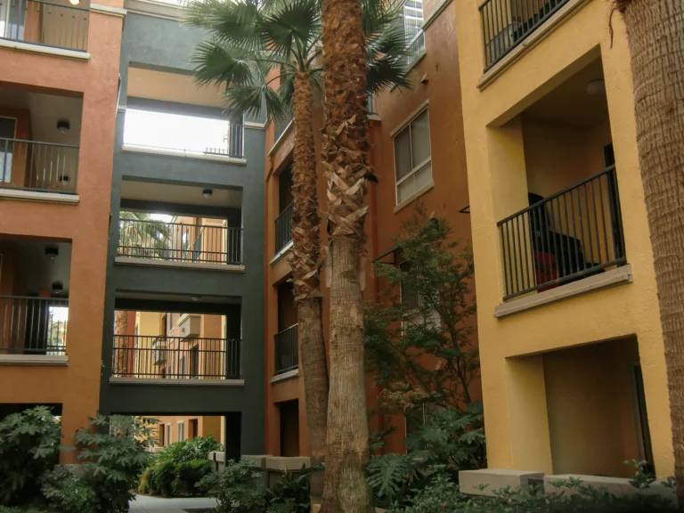 Apartment building courtyard with orange and yellow walls, balconies, tall palm trees, and lush green plants. Multiple floors and railings are visible, creating an inviting and open outdoor space.