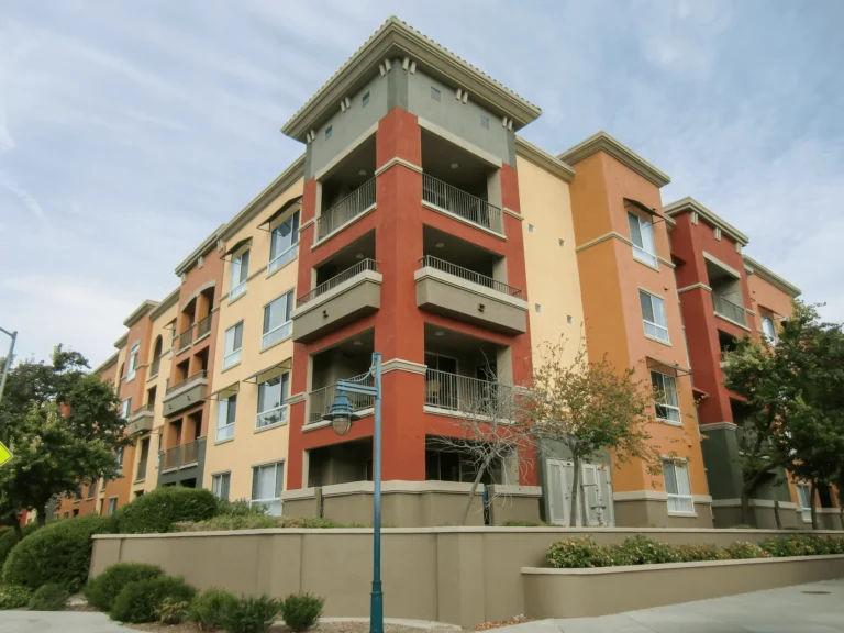 A modern four-story apartment building with red, orange, and beige exterior walls, balconies, and large windows, surrounded by trees and shrubs under a cloudy sky.