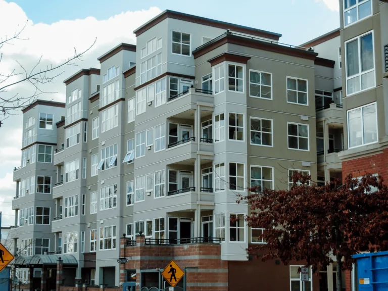A modern, multi-story apartment building with large windows and balconies, featuring beige and white exterior walls. A pedestrian crossing sign and a tree with brown leaves are visible in the foreground.