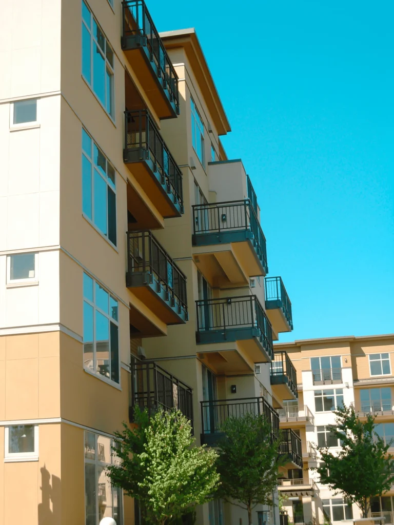 Modern apartment building with beige and white walls, large windows, and several balconies with black railings. Green trees line the front of the building, and the sky is clear and blue.