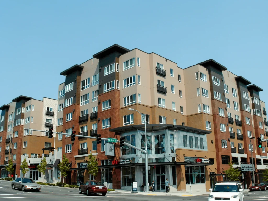 A modern multi-story apartment building with shops on the ground floor stands at a busy street corner. Cars wait at a traffic light, and pedestrians walk on the sidewalks under a clear blue sky.
