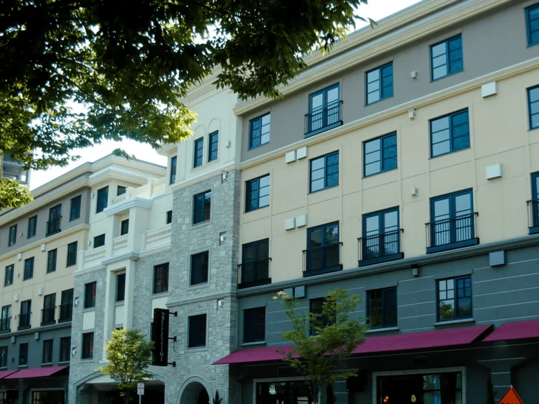 A modern four-story apartment building with large windows, a mix of stone and stucco exterior, and red awnings over the ground-floor shops, framed by green trees in the foreground.