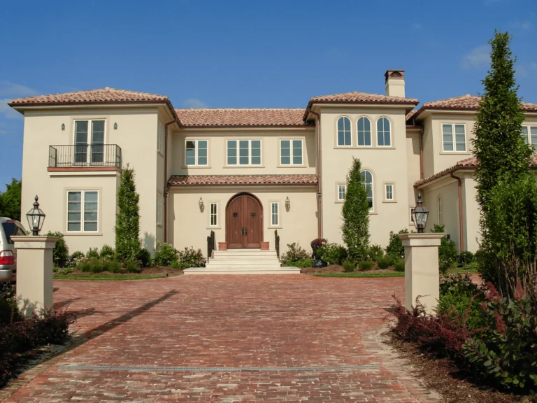 Large beige Mediterranean-style house with a red-tile roof, arched wooden front doors, multiple windows, balconies, and a spacious brick driveway lined with greenery under a clear blue sky.
