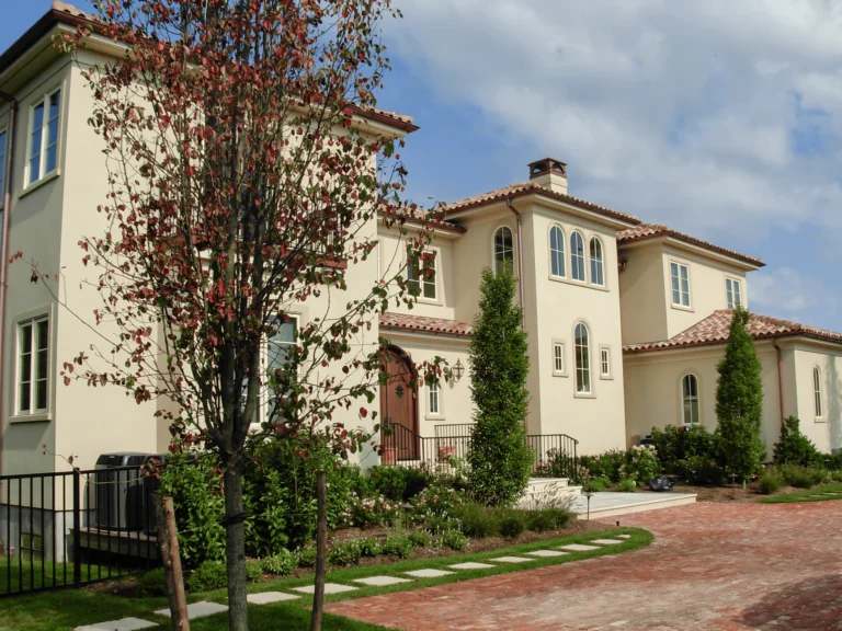 A large, beige, two-story house with red-tiled roof, arched windows, and a wooden front door sits behind a tree and landscaped garden, with a brick driveway leading to the entrance under a partly cloudy sky.