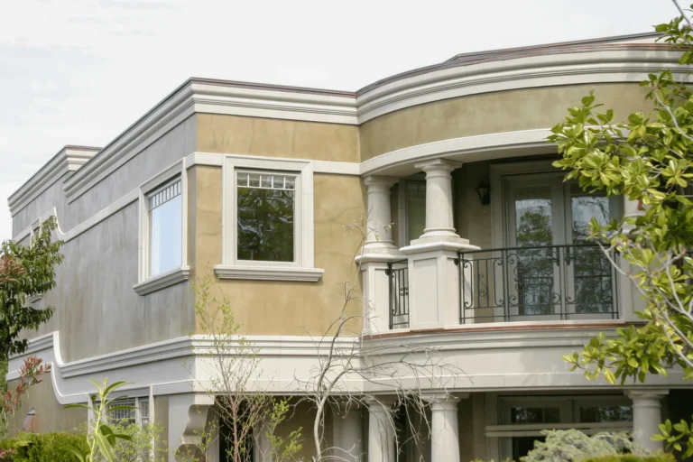 Two-story modern house with tan stucco walls, large windows, white trim, and a small balcony with columns and a black metal railing. Trees and greenery are visible in the foreground.