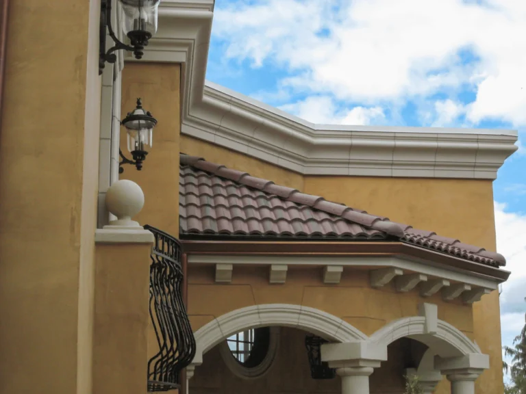 A close-up of a Mediterranean-style building with yellow stucco walls, red clay roof tiles, white trim, decorative wrought iron balcony, and classic lanterns under a partly cloudy blue sky.