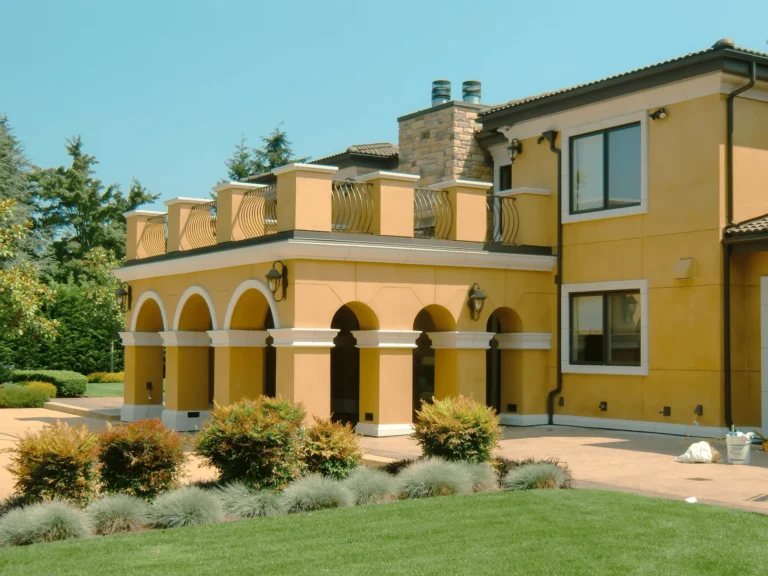 A large yellow stucco house with arched columns and a second-floor terrace, surrounded by manicured bushes, greenery, and a well-kept lawn on a sunny day.