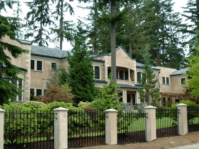 A large, beige two-story house with a black metal fence, surrounded by tall trees and lush green landscaping, sits on a spacious lot.