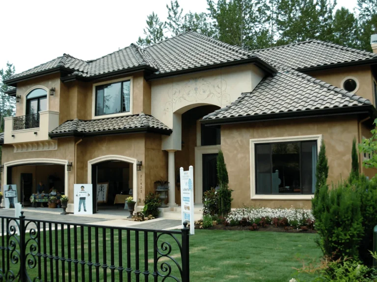 Two-story modern stucco house with a tile roof, manicured lawn, and decorative plants. Open doors display interior, and signs in front suggest the house is for sale or part of an open house event.