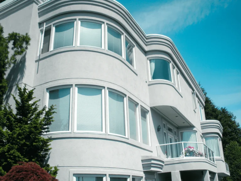 Modern two-story house with curved white exterior, large windows, and a small balcony decorated with flowers, surrounded by green trees under a clear blue sky.