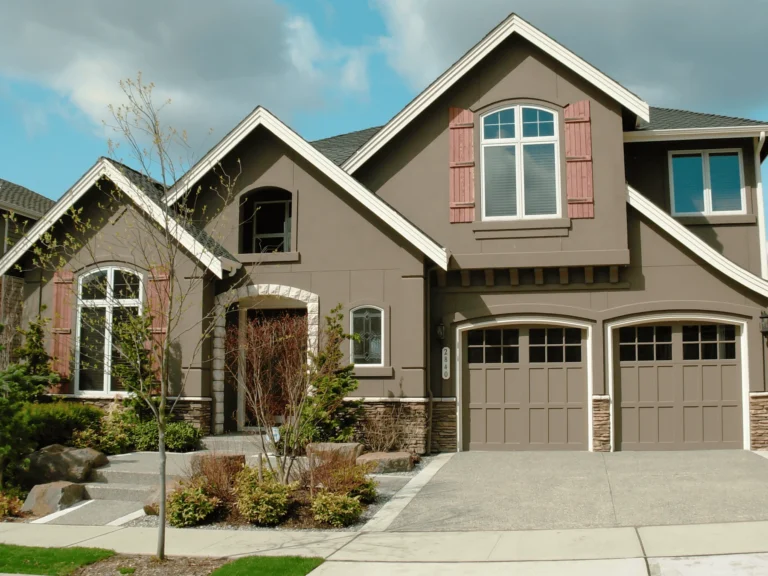 Two-story suburban house with brown siding, stone accents, two garage doors, and shuttered windows. A small tree and shrubs are in the landscaped front yard under a partly cloudy sky.