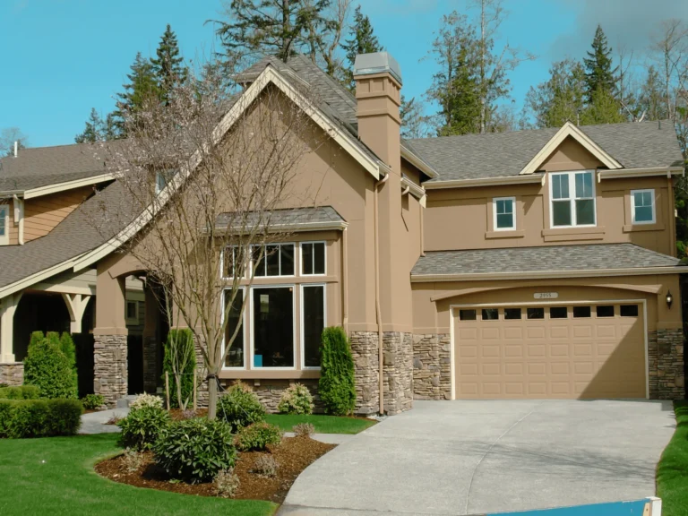 A modern two-story house with beige walls, stone accents, large windows, and a double garage, surrounded by trimmed bushes, trees, and a neatly maintained lawn under a clear blue sky.