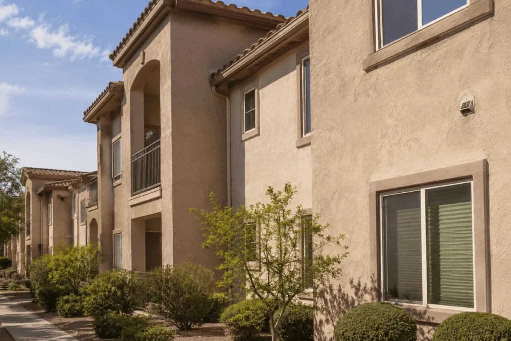 Two-story beige apartment building with outdoor balconies, large windows, and tiled roofs, surrounded by small bushes and trees under a partly cloudy blue sky.