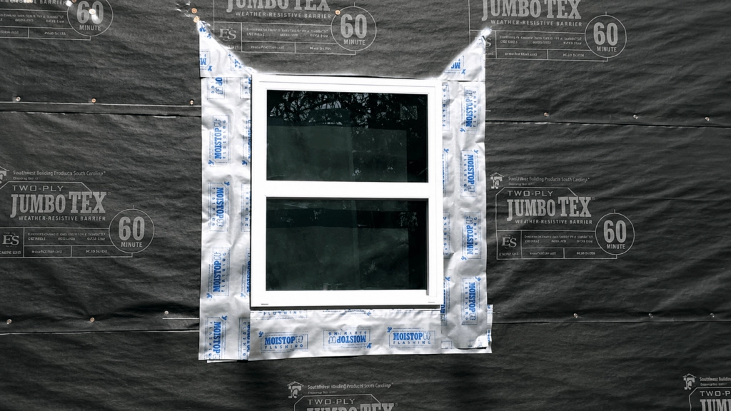 A white-framed window is installed in a wall covered with black building wrap. The window is surrounded by silver flashing tape for waterproofing, and trees are reflected in the window glass.