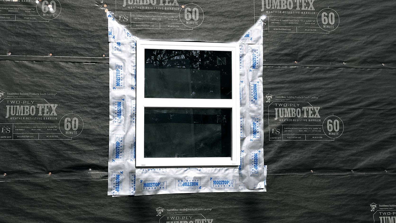 A white-framed window is installed in a wall covered with black building wrap. The window is surrounded by silver flashing tape for waterproofing, and trees are reflected in the window glass.