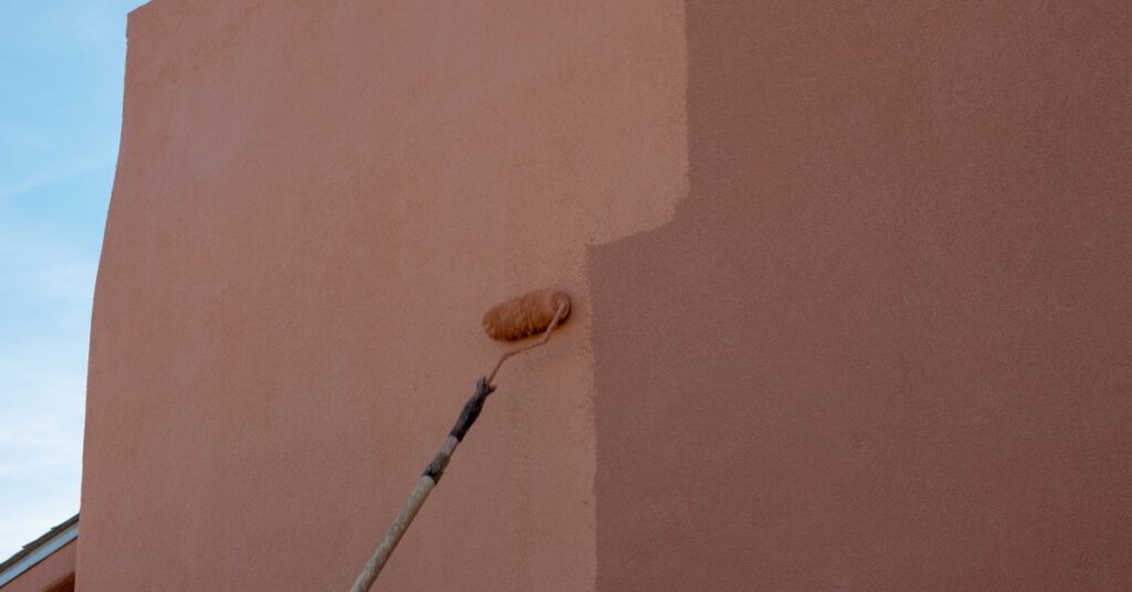 Someone is painting a large Adobe house with a painting roller brush. The brush extends and the end is brown with paint.