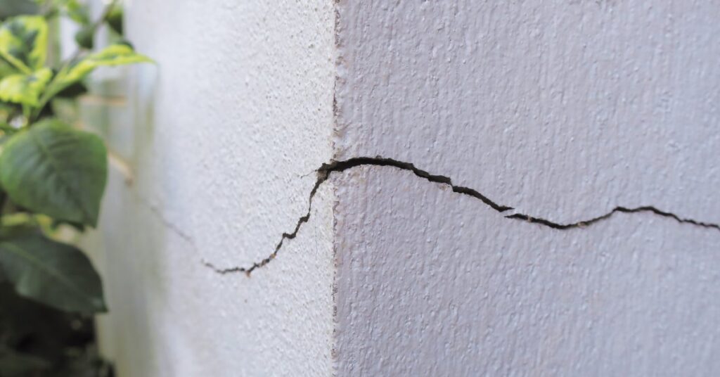 A horizontal, curved crack on an exterior corner of a building's white wall. Green plants are in the background.