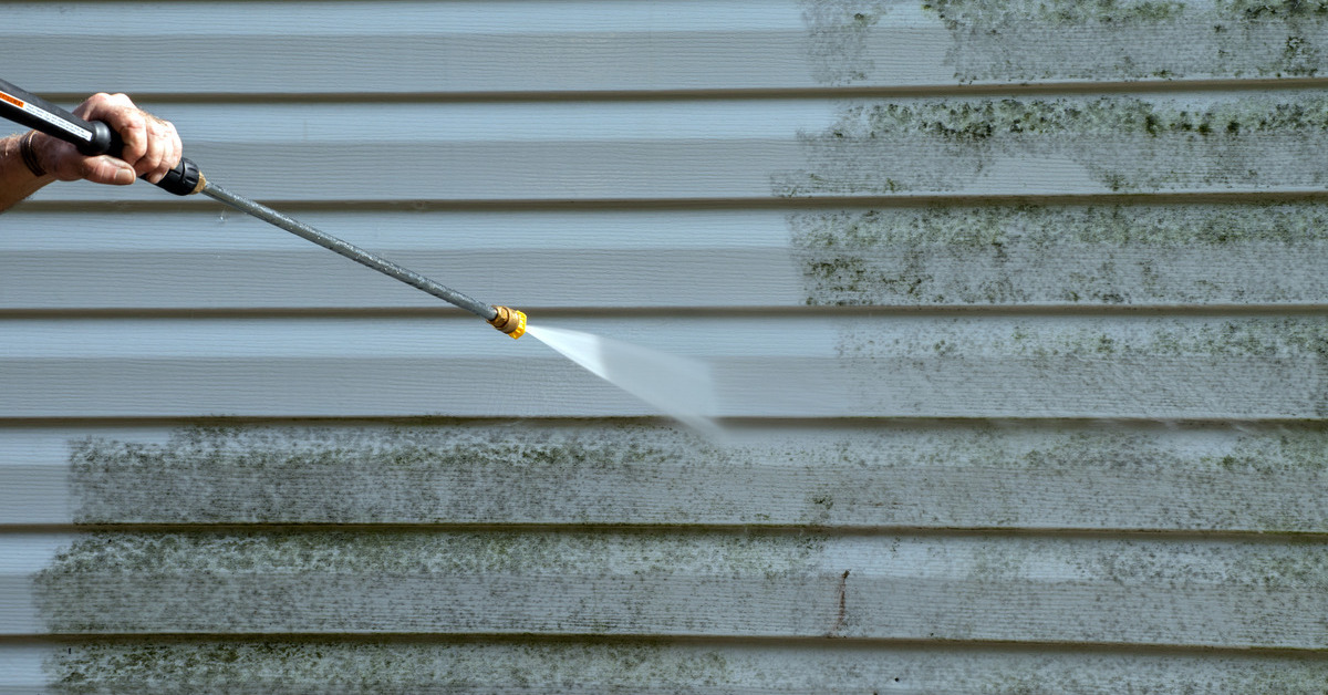 A person holding a pressure washer removes black mold from white vinyl siding using water on a home's exterior.