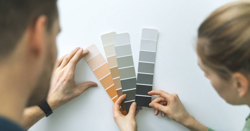 A man and a woman hold up paint samples against a wall. The shades include orange, brown, and gray.